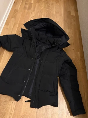 Svart pufferjacka från Canada Goose - Säljer en svart pufferjacka från Canada Goose med huva och dragkedja framtill. Jackan har en stilren look med diskreta knappar och rejäla fickor. Perfekt för kalla dagar när du vill hålla värmen och ändå se snygg ut. Äkta så klart kvitton finns och kan tänka mig ner i pris vid snabbäffar kan även träffas upp 