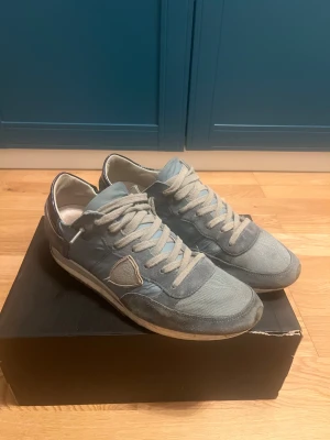 Blå Philippe Model sneakers  - Snygga blå sneakers från Philippe Model med grå detaljer. Skorna har ovandel i mocka och textil, klassisk snörning och den ikoniska sköld-loggan på sidan. Perfekta för dig som gillar en stilren och sportig look. Perfekt till vintern 