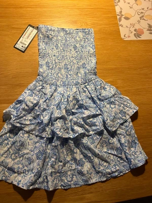 Blåvit axelbandslös kortklänning PB Sister - Supersöt axelbandslös kortklänning från PB Sister i blått och vitt med blommigt mönster. Klänningen har smockad överdel och volangig kjol i två lager. Perfekt för sommarens varma dagar och ger en fräsch och trendig look.