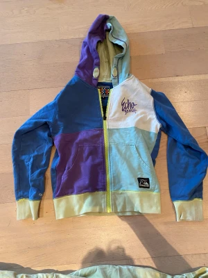 Färgglad hoodie från Quiksilver - Säljer en unik hoodie från Quiksilver med coola färgblock i blått, lila, vitt och ljusgrönt. Tröjan har huva med snörning, dragkedja framtill och två fickor. Broderad Echo Beach-logga på bröstet och Quiksilver-märke vid fickan. Perfekt för dig som gillar streetwear och färg!