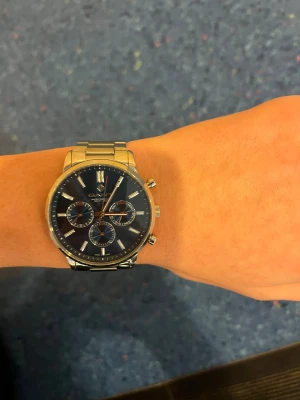GANT herrklocka med blå urtavla - Stilren herrklocka från GANT med blå urtavla och silverfärgat armband i rostfritt stål. Klockan har tre mindre urtavlor, kronograf och tydliga indexmarkeringar. Klassisk rund form och robust länkarmband som ger en modern look. Har tyvärr ingen box eller kvitto.