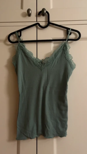 Spetslinne - Snygg grön/turkos linnetopp med spetsdetaljer längs v-ringad halsringning. Tunn och mjuk, med smala axelband och figurnära passform. Perfekt för att skapa en chill och trendig look. Storleken är xs/s/m. ALDRIG ANVÄND, som ny!!
