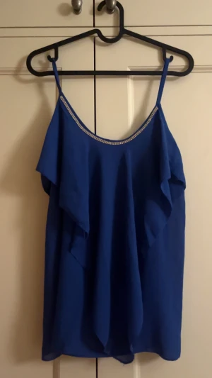 Blå volangtopp med detaljer - Snygg blå linnetopp med tunna axelband och diamantdetaljer längs halsringningen. Toppen har ett flowigt fall med volang framtill som ger en cool och feminin vibe. Perfekt för dig som vill sticka ut med lite extra bling och färg.