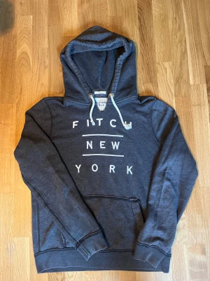 Abercrombie & Fitch hoodie  - Snygg mörkgrå hoodie från Abercrombie & Fitch med vit text 'FITCH NEW YORK' på bröstet. Tröjan har huva med snörning, ribbade muddar och klassisk känguruficka framtill. Perfekt för chill dagar och streetstyle.