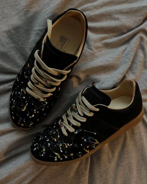 Svarta sneakers inspirerad av GATS - Unika svarta sneakers med coola färgstänk i gult, blått, rött och vitt över hela ovandelen. Skorna har beige snörning, rund tå och platt gummisula. Materialet är mocka och skinn, vilket ger en lyxig känsla och snygg kontrast. Perfekt för dig som vill sticka ut med din stil. Inspirerade av GATS. Helt nya.