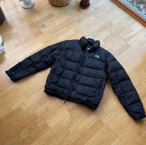 Svart pufferjacka från The North Face - Svart pufferjacka från The North Face i storlek M. Jackan har en hög krage, två sidofickor med dragkedja och en klassisk logga på bröstet. Materialet är syntet och dun, perfekt för kalla dagar. ❄️  Skick 8/10, jätte fint skick, bara defekt på fickans dragkedja ✅