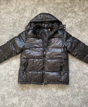 Svart pufferjacka från Polo Ralph Lauren - Svart glansig pufferjacka från Polo Ralph Lauren med huva och dragkedja framtill. Jackan har två fickor med dragkedja och klassisk röd logga på bröstet. Perfekt för kalla dagar och har en modern, boxig passform.