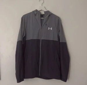 Grå vindjacka från Under Armour - Säljer en snygg tvåfärgad vindjacka från Under Armour i mörkgrått och ljusgrått. Jackan har huva, dragkedja framtill och diskret logga på bröstet. Perfekt för blåsiga dagar och sportiga outfits.