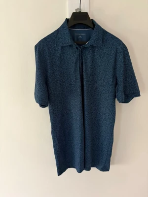 Blå mönstrad golfpiké Galvin Green - Snygg blå skjorta från Galvin Green med diskret mönster och klassisk krage. Skjortan har korta ärmar och en stilren look som passar perfekt till sommaren. Tillverkad i ett lätt och bekvämt material som andas. Storlek: L passar M. Nypris: 999kr