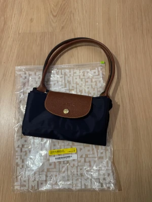 Mörkblå Longchamp handväska - Snygg mörkblå handväska från Longchamp med bruna skinnhandtag och lock. Väskan har en klassisk, stilren design med guldfärgad knapp och är tillverkad i slitstark polyester. Perfekt storlek för att få med allt du behöver och passar till många olika looks.
