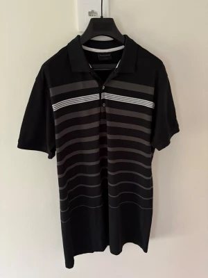 Svart randig golfpiké Galvin Green - Svart golfpiké från Galvin Green med grå och vita horisontella ränder. Klassisk krage och knappslå framtill, korta ärmar och mjukt material som känns skönt mot huden. Perfekt för dig som gillar stilren design med sportig touch. Storlek: L passar M. Nypris: 999kr
