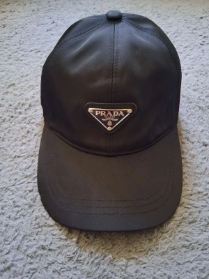 Svart keps från Prada i nylon - Snygg svart keps från Prada i återvunnen nylon. Klassisk modell med böjd skärm och knapp på toppen. Perfekt accessoar för en stilren look och enkel att matcha med olika outfits.