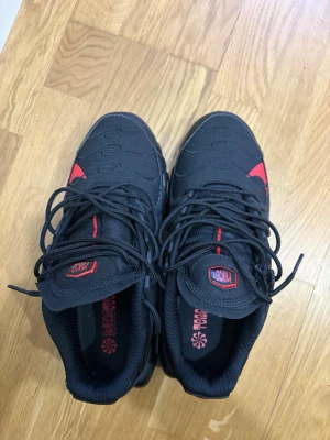 Svarta Nike Air Max Tn Plus sneakers - Snygga Nike Air Max Tn Plus sneakers i svart med röda detaljer och tydlig Nike-logga på. Skorna har en kraftig sula och är tillverkade i textil och syntetmaterial. Perfekta för dig som gillar streetwear och vill sticka ut.