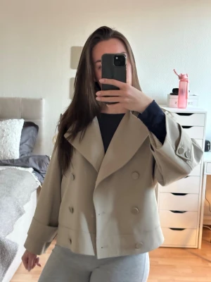 Kort beige trenchcoat  - ❗️ Villig att sänka priset❗️ Stilren kort beige trenchcoat med dubbelknäppning och bred krage. Jackan har vida ärmar, knappar och resår i ryggen för en snygg passform. Bild 3 visar färgen bäst! Använd kanske 3-4 gånger bara tyvärr men den kan få någon som använder den oftare 💕💕Det är storlek XS men skulle säga att den är mer som en S.  Nypris 1750kr