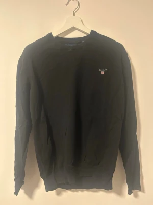 Svart stickad tröja från GANT - Klassisk svart tröjafrån GANT med rund halsringning och diskret logga broderad på bröstet. Tröjan har långa ärmar och ribbade muddar vid ärmslut och nederkant. Tröjan är in storlek M. Pris kan diskuteras. Fler bilder skickas vid behov.