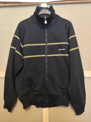 Adidas Originals track jacket West Germany vintage svart/gul M - Vintage adidas Originals track jacket producerad av DESCENTE under adidas-licens. Märkt BRD / W. Germany – detaljer som förknippas med äldre och mer eftertraktade adidas-plagg med stark kultstatus. Känd för hög kvalitet, bättre passform och tidlös design. En riktig raritet och samlarobjekt. Svart med gula och vita linjer, trefoil-logo och original dragkedja. Mycket fint begagnat skick. Storlek M.