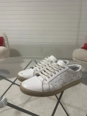 Ysl Skor - Snygga vita sneakers med silverfärgade stjärnor på sidorna och vita snören. Skorna har en klassisk låg profil och en beige sula. Perfekta för dig som gillar en clean look med lite extra detaljer.