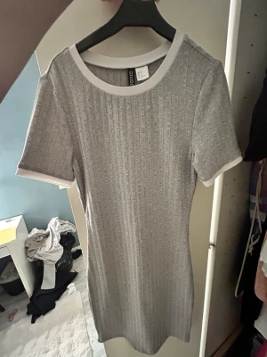 Grå ribbad klänning från Divided - Kortärmad grå ribbad klänning från Divided med vit kant vid halsringning och ärmslut. Klänningen har en kroppsnära passform och är tillverkad i ett mjukt, stretchigt material som ger en bekväm känsla. Perfekt för dig som gillar en enkel men stilren look.