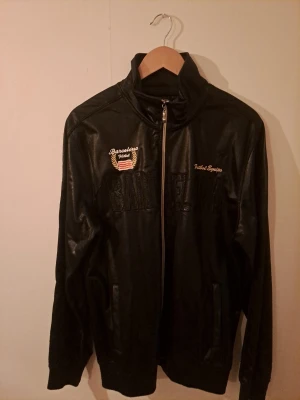 BARCELONA ZIPPER  med dragkedja - Snygg svart jacka med glansig finish och broderad text 'Barcelona' samt detaljer i guld och rött. Jackan har hög krage och hel dragkedja framtill. Perfekt för dig som gillar sportig stil och vill sticka ut med en unik jacka.