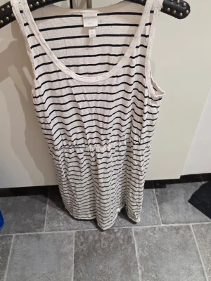 Randig vit klänning från H&M - Säljer en vit ärmlös klänning från H&M med svarta horisontella ränder. Klänningen har rund halsringning, resår i midjan och är tillverkad i mjuk bomull. Perfekt för varma dagar och enkel att styla med sneakers eller sandaler.