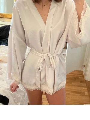 Ljusrosa kort morgonrock med spets - Supersöt ljusrosa morgonrock i kort modell med knytband i midjan och fina spetsdetaljer vid ärmslut och nederkant. Tillverkad i ett mjukt, glansigt material som känns lyxigt mot huden. Perfekt för mysiga hemmakvällar.