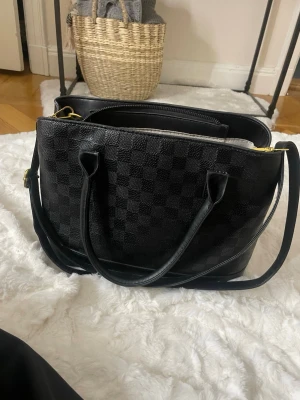 Svart rutmönstrad handväska - Snygg svart handväska med klassiskt rutmönster och dubbla handtag. Väskan har även en lång axelrem och detaljer i guld. Tillverkad i ett tåligt konstlädermaterial med struktur. Perfekt för dig som gillar stilrena accessoarer med lyxig känsla.