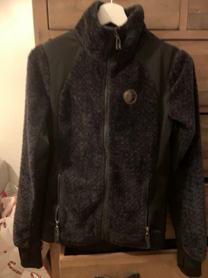 Ridkläder - svart teddyjacka  - Mysig svart teddyjacka med hög krage och hel dragkedja framtill. Jackan har två fickor med dragkedja och detaljer i slätare tyg på axlar och sidor. Använd men i bra skick.