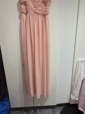Ljusrosa långklänning med draperad överdel - Elegant ljusrosa långklänning med smala axelband och vackert draperad överdel. Klänningen har en flowy kjol och en romantisk känsla, perfekt för speciella tillfällen. Tillverkad i ett lätt och mjukt material som faller snyggt.