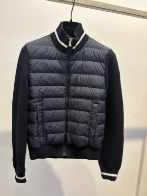  Moncler Cardigan (14Y) -  Moncler Cardigan med quiltad front och stickade ärmar. Jackan har dragkedja framtill, ribbade muddar och krage med vita ränder. Moncler-logga på ärmen ger en exklusiv touch. Perfekt för dig som vill ha både stil och komfort  Använd någon gång, väldigt bra skick stl 164 (14Y)köpt i Londonbutiken 2025 Liten i storlek så motsvarar 152-158  Önskar man påsen från butiken går den att få vid upphämtning ( skickas ej) 
