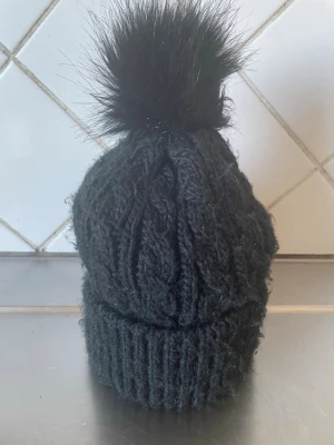 Svart stickad mössa med pälsboll H&M - Snygg svart stickad mössa från H&M med grovt flätmönster och stor fluffig pälsboll på toppen. Perfekt för kalla dagar när du vill hålla stilen. Mössan är mjuk och har ribbad kant för extra komfort.