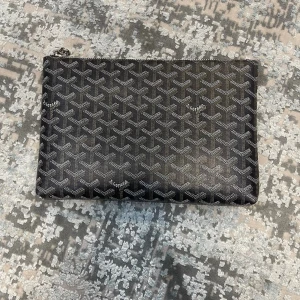 Svart väska med mönster - Snygg svart kuvertväska med deras ikoniska vita mönster. Väskan har en rektangulär form och dragkedja på baksidan. Tillverkad i canvas med detaljer i läder. Perfekt för att bära det viktigaste med stil.