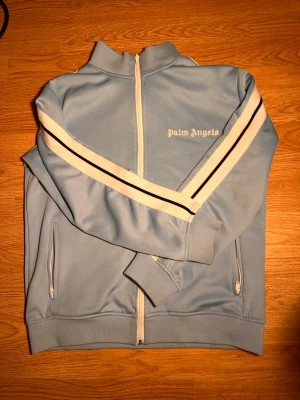 Palm Angels blå träningsjacka - Snygg ljusblå träningsjacka från Palm Angels med vita och svarta ränder längs ärmarna. Jackan har dragkedja framtill, ståkrage och två fickor med dragkedja. Märkets logga är broderad på bröstet. Perfekt för en sportig och trendig look.