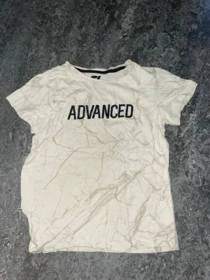 Vit t-shirt med tryck 'ADVANCED' - Vit t-shirt med grått marmorerat mönster och svart texten 'ADVANCED' på bröstet. Klassisk rund halsringning och korta ärmar. Perfekt för en avslappnad och trendig stil.