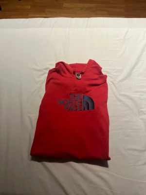 Röd hoodie från The North Face - Snygg röd hoodie från The North Face med stor logga broderad i blått på bröstet. Klassisk känguruficka framtill och huva. Perfekt för dig som vill ha en stilren och bekväm hoodie med streetwear-vibe. Tillverkad i mjukt bomullsmaterial.