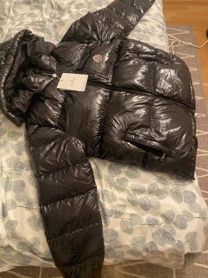 Svart glansig dunjacka från Moncler - Snygg svart dunjacka från Moncler med glansig finish och klassisk logotyp på bröstet. Jackan har huva, dragkedja framtill och praktiska fickor med dragkedja. Perfekt för kalla vinterdagar och riktigt trendig look.
