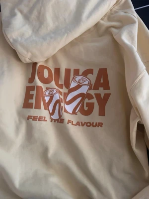 Gul hoodie från Jolucca Energy - Ljusgul hoodie från Jolucca Energy med tryck på ryggen i orange och vitt, samt liten broderad logga framtill. Hoodien har huva och känguruficka. Materialet är mjukt bomullstyg, perfekt för en chill och avslappnad stil.