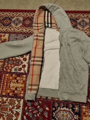 Grå hoodie från Burberry med rutig huva - Säljer en grå hoodie från Burberry med den klassiska beige-rutiga insidan i huvan. Hoodien har dragkedja, fickor framtill och ribbade muddar. Materialet är mjukt och skönt, perfekt för chill dagar. Ikonisk Burberry-stil på huvan för extra lyxig känsla.