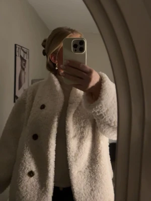 Vit teddyjacka med knappar - Supermysig vit teddyjacka med stora svarta knappar framtill. Jackan har en rak passform och är gjord i fluffigt teddy-material som ger en trendig och avslappnad look. Perfekt för att hålla sig varm och snygg under kyliga dagar.