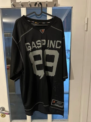 Oversized svart mesh t-shirt GASP INC - Säljer en oversized svart mesh t-shirt från GASP INC med tryckta siffror 89 fram och bak. T-shirten har korta ärmar, sportig look och är tillverkad i luftig polyester. Perfekt för dig som gillar streetwear eller gymstyle.