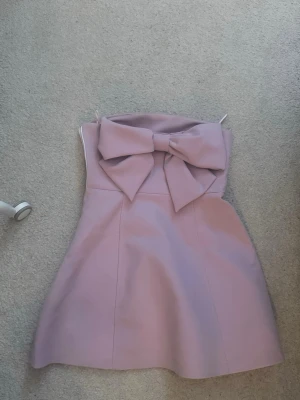 Rosa klänning med rosett - Supersöt ljusrosa axelbandslös klänning med stor rosett i ryggen. Kort modell med figurnära passform upptill och lätt utställd kjol. Perfekt för sommarens alla tillfällen. Materialet känns slätt och stadigt. använt en gång. men behövt sy om dragkedjan därav en vit, men den syns ej så mycket! 
