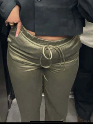 Olivgröna mjukisbyxor med dragsko - Snygga olivgröna mjukisbyxor med rak passform och resår i midjan. Byxorna har dragsko och sidofickor, tillverkade i ett mjukt och glansigt material som ger en chill och trendig vibe.