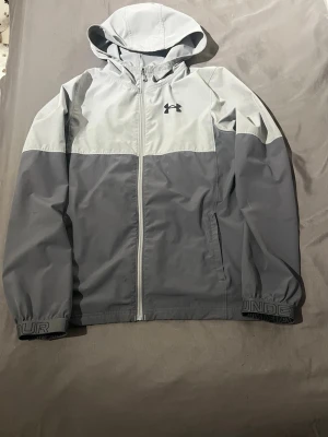 Grå vindjacka från Under Armour - Snygg tvåtonad grå vindjacka från Under Armour med huva och dragkedja framtill. Jackan har elastiska muddar med logotypdetaljer och två sidofickor. Perfekt för blåsiga dagar och sportiga outfits. Materialet är lätt och vindtätt.