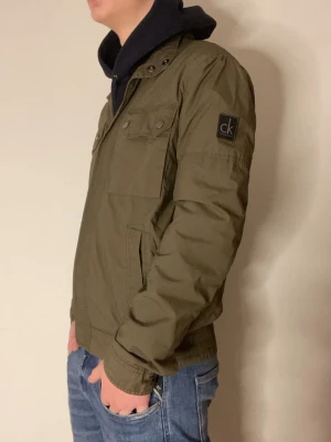 Olivgrön jacka från Calvin Klein - Snygg olivgrön jacka från Calvin Klein med två bröstfickor med lock och knappar, samt sidofickor. Jackan är ganska tunn och kan användas som en vind jacka. Modellen är 178cm!