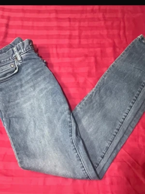 Blå raka jeans med knappar - Snygga blå jeans med raka ben och klassisk femficksdesign. Jeansen har knäppning med två synliga metallknappar och bälteshällor. Materialet är denim i en tvättad blå nyans med lätt slitning för en avslappnad look.