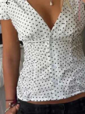 Vit prickig blus med v-ringning - Säljer en söt vit blus med svarta prickar och v-ringning. Blusen har korta ärmar, knappar framtill och figurnära passform. Perfekt till jeans eller kjol för en trendig look.(inte min bild)