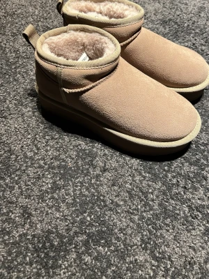 Beige fluffiga slip-in tofflor - Säljer ett par beige slip-in tofflor med mjukt, fluffigt foder och ovandel i mockaimitation. De har en rejäl sula i gummi och en låg ankelhöjd. Perfekta för att hålla fötterna varma och bekväma hemma eller ute.