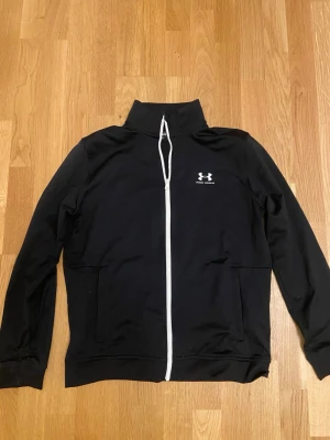 Svart Under Armour Zip-tröja - Säljer en stilren svart zip-tröja från Under Armour med vita detaljer. Tröjan är i mycket fint skick och har endast använts som vardagsplagg vid ett fåtal tillfällen. Nypris: 700kr
