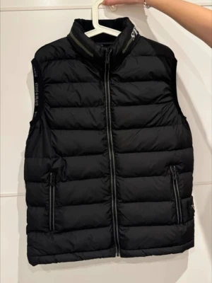 Svart dunväst från S.I med patch - Snygg svart dunväst med hög krage och dragkedja framtill. Västen har två fickor med dragkedja och logotypdetalj på kragen samt S.I patch. Klassisk quiltad design och perfekt för lager-på-lager. Materialet är dunfyllning och yttertyg i nylon. Köpt på Vinted men den satt ej bra på mig.