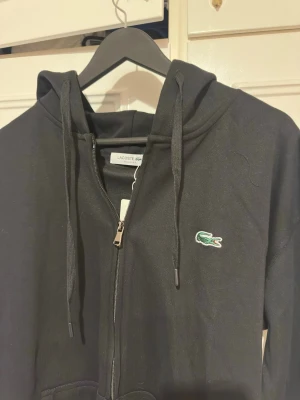 Svart zip hoodie från Lacoste - Snygg svart hoodie från Lacoste med dragkedja framtill och klassisk krokodil-logga på bröstet. Modellen har huva med dragsko och fickor framtill. Regular fit och tillverkad i mjukt bomullsmaterial för skön känsla. Perfekt för en avslappnad och stilren look.