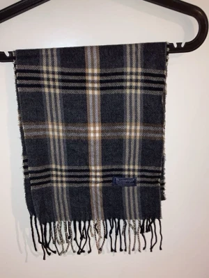 Rutig halsduk från Burberrys - Säljer en klassisk rutig halsduk från Burberrys i mörkgrå och beige toner. Halsduken är tillverkad i 100% lammull och har fransar längs nederkanten. Perfekt för detta kalla väder.